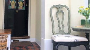 Una puerta negra con un vitral, una silla verde con cojín, y una mesita con un jarrón de flores amarillas.