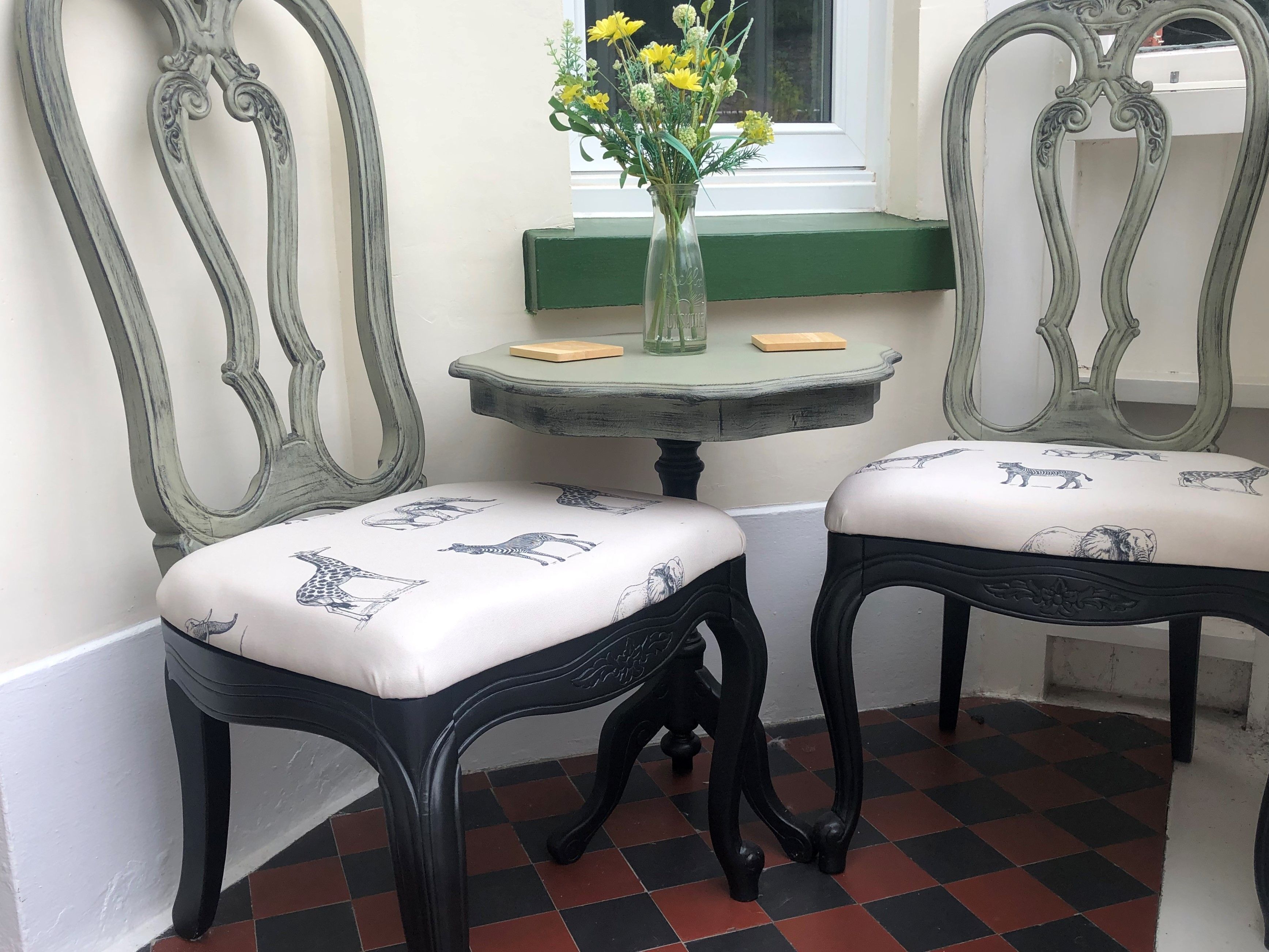 Two black chairs with white cushions featuring animal prints, a small round table with a vase of yellow flowers, and a window with a green frame.