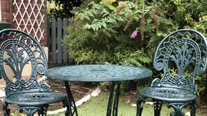 Una mesa metálica redonda con dos sillas, rodeada por un jardín con césped verde y diversas plantas.
