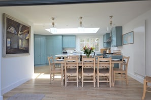 A spacious dining area with a large table and chairs, a kitchen in the background, and a skylight above.