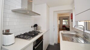 A modern kitchen with white tiles, a gas stove, a toaster, and a stainless steel sink. - Pebble Cottage (Ventnor)
