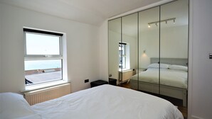 A bedroom with a large window, a bed, a wardrobe with a mirror, and a radiator. - Pebble Cottage (Ventnor)