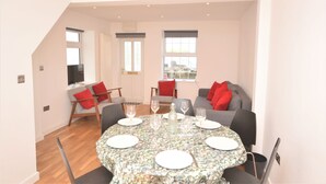 A dining area with a round table set for four, surrounded by chairs. There is a sofa with red cushions in the background. - Pebble Cottage (Ventnor)