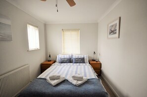 A neatly made bed with striped bedding, two bedside tables with lamps, a window with blinds, and a framed picture on the wall. - Harbour View (East Cowes)