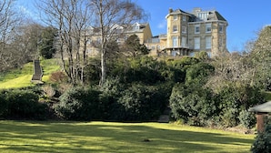 A large, historic building with multiple stories and a turret, surrounded by lush greenery and a well-maintained lawn.