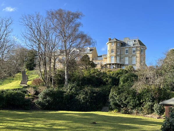 A large, historic building with multiple stories and a turret, surrounded by lush greenery and a well-maintained lawn.