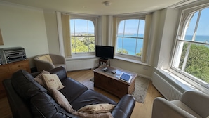 A living room with a leather sofa, a wooden coffee table, and a television. There are large windows with a view of the sea and greenery.