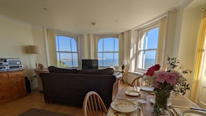 A dining area with a table set for two, a sofa, and a view of the sea through large windows.