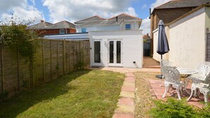 Un jardin à l’arrière de la maison, avec une terrasse blanche, une clôture en bois et un petit espace aménagé en jardin.