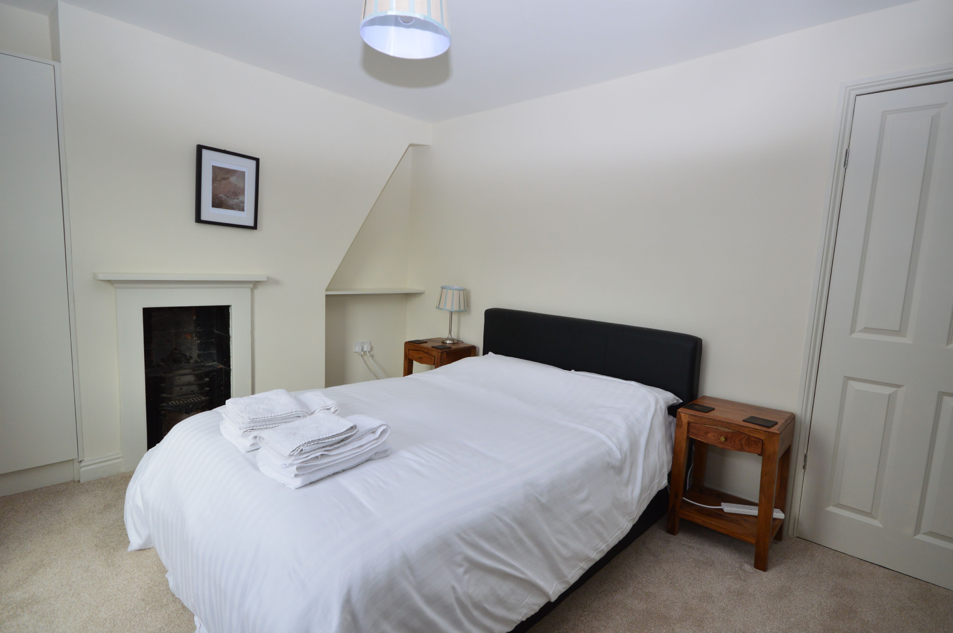 A bedroom with a bed, a nightstand, a lamp, a framed picture, and a fireplace.
