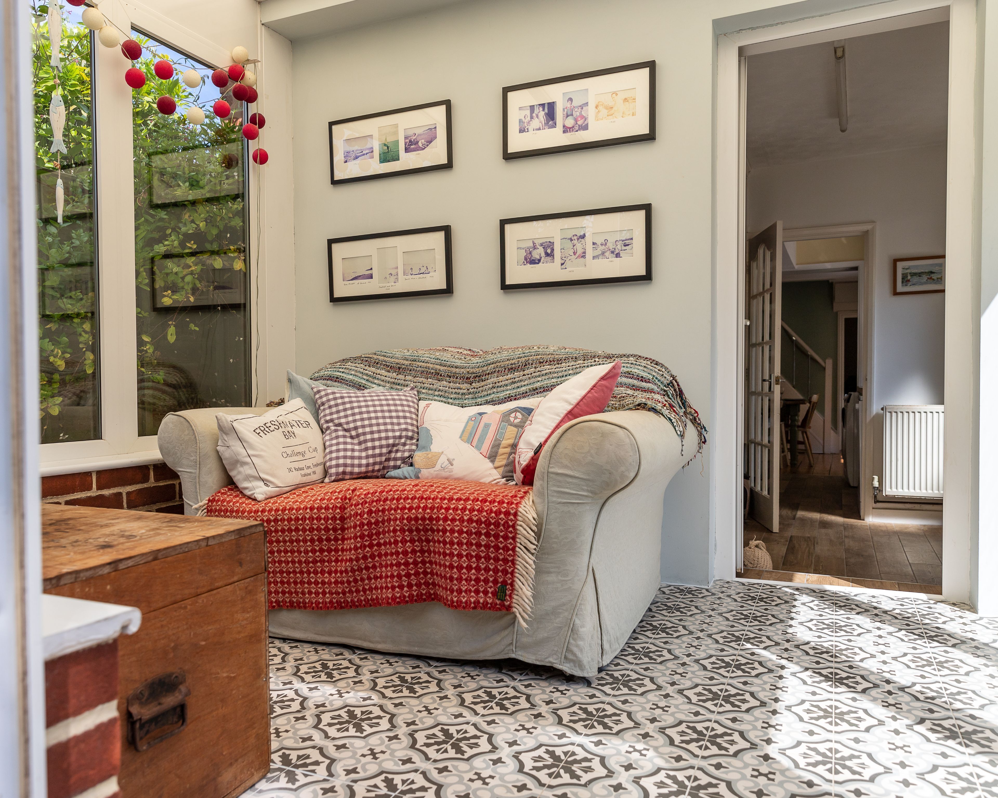 A cozy living room with a patterned floor, a grey sofa with multiple cushions, and framed pictures on the wall.