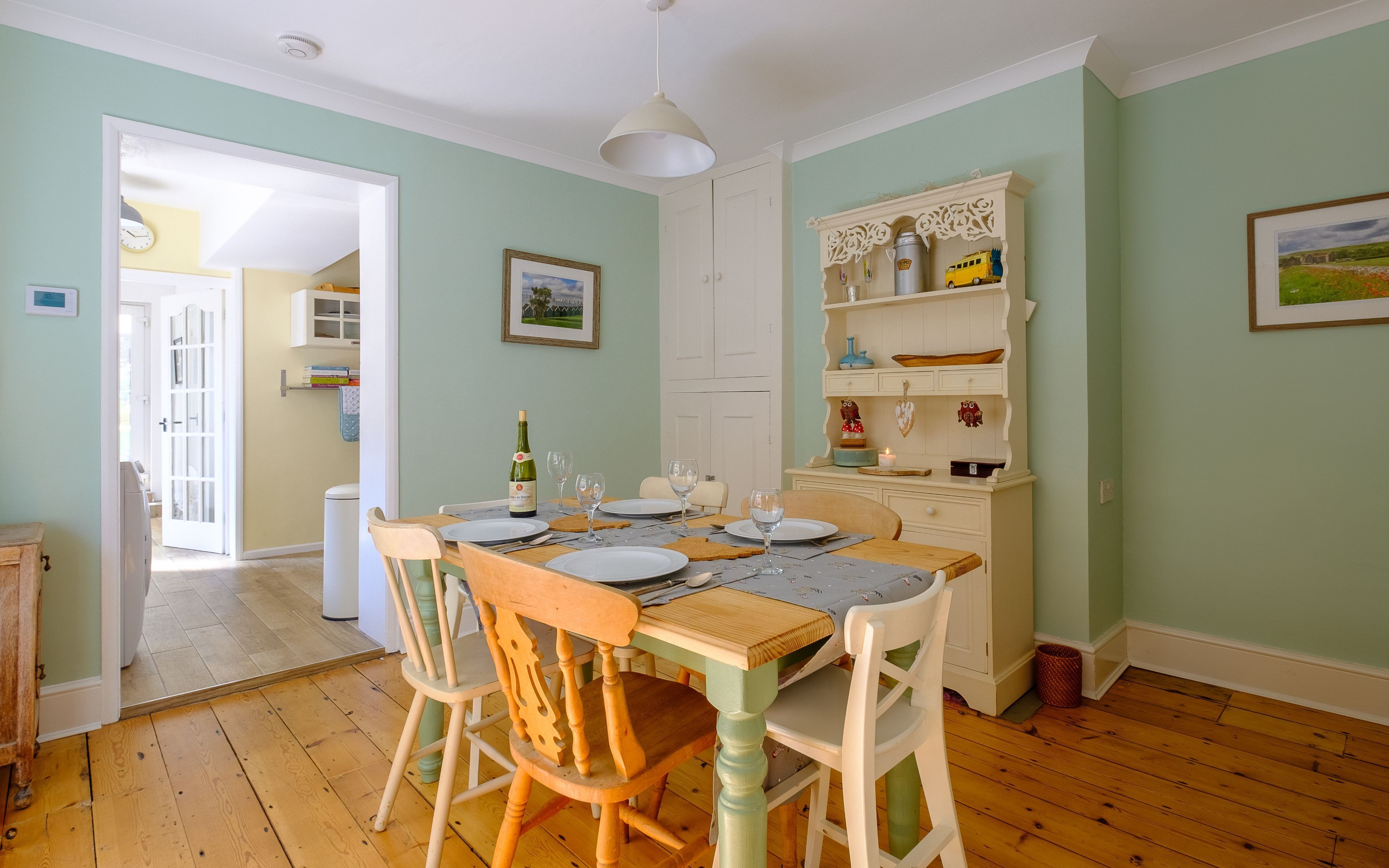A dining area with a wooden table set for four, surrounded by chairs. A white cabinet with decorative items is to the right.