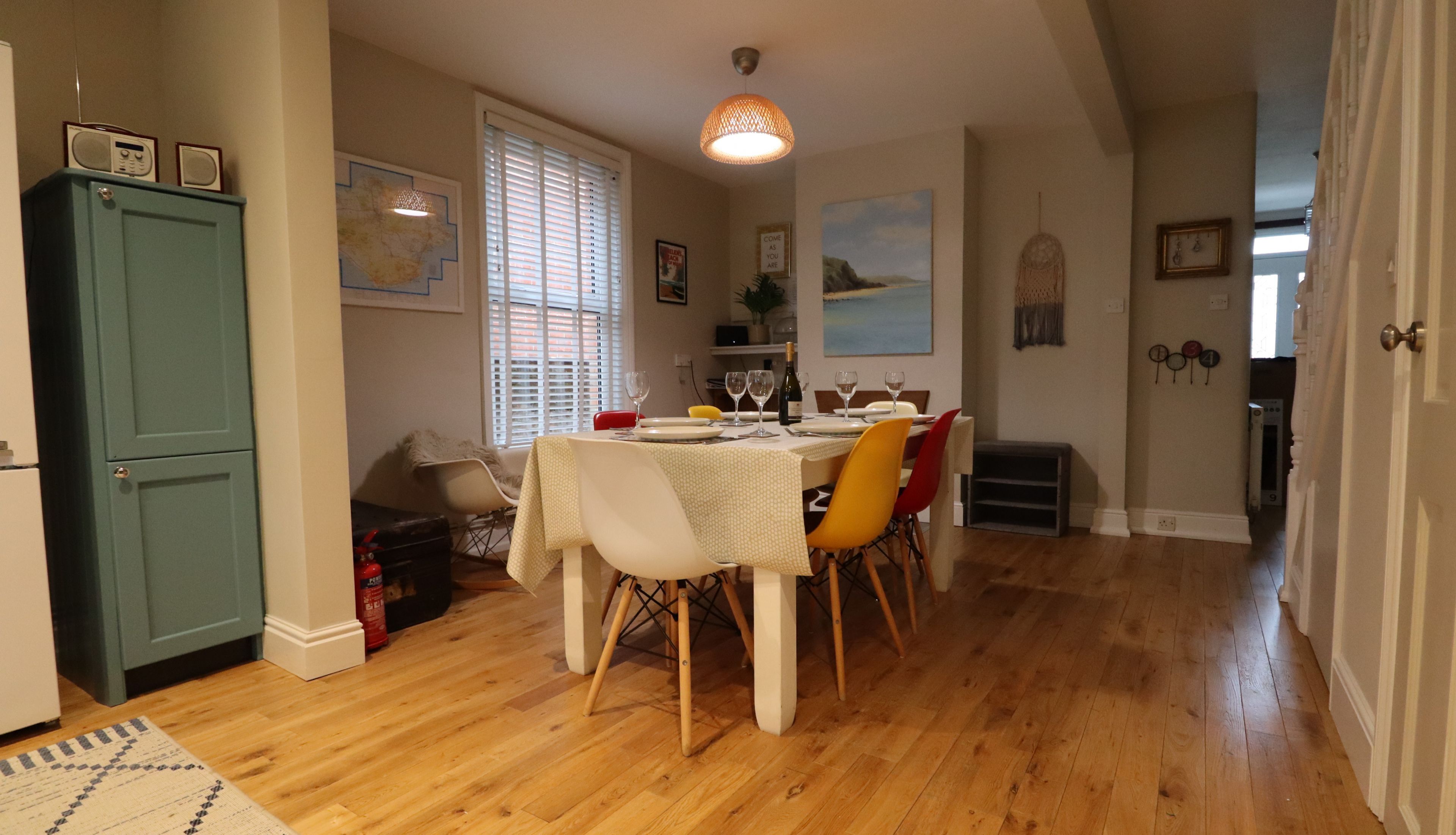 A dining area with a wooden table set for four, surrounded by chairs. There is a green cabinet to the left and a window with blinds.