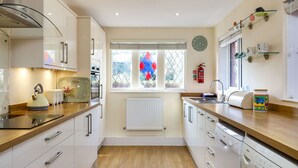 A modern kitchen with white cabinetry, a built-in oven, a dishwasher, and a window with diamond-patterned glass. - Mole Cottage (Shanklin)