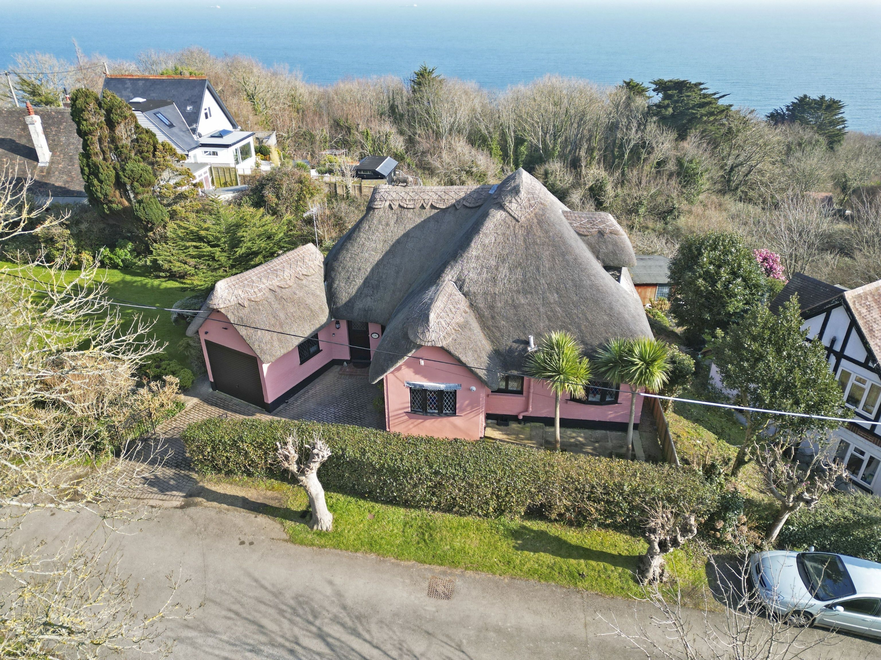A house with a thatched roof and a pink exterior, surrounded by a well-maintained garden and a view of the sea.