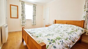 A bedroom with a wooden bed, a bedside table, a mirror, and a window with floral curtains. - The Willows (Newport)