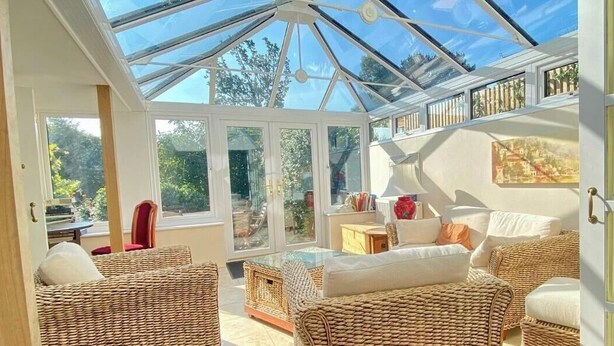 Una habitación iluminada por el sol con muebles de mimbre, techo de cristal y vistas al verde exterior.