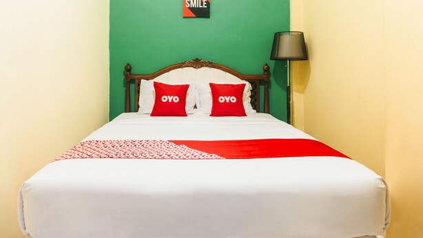 Un lit recouvert d’une couvre-lit blanche et rouge, deux oreillers rouges ornés du mot « OYO », une tête de lit en bois et un mur vert sur lequel est affiché le mot « SMILE ».