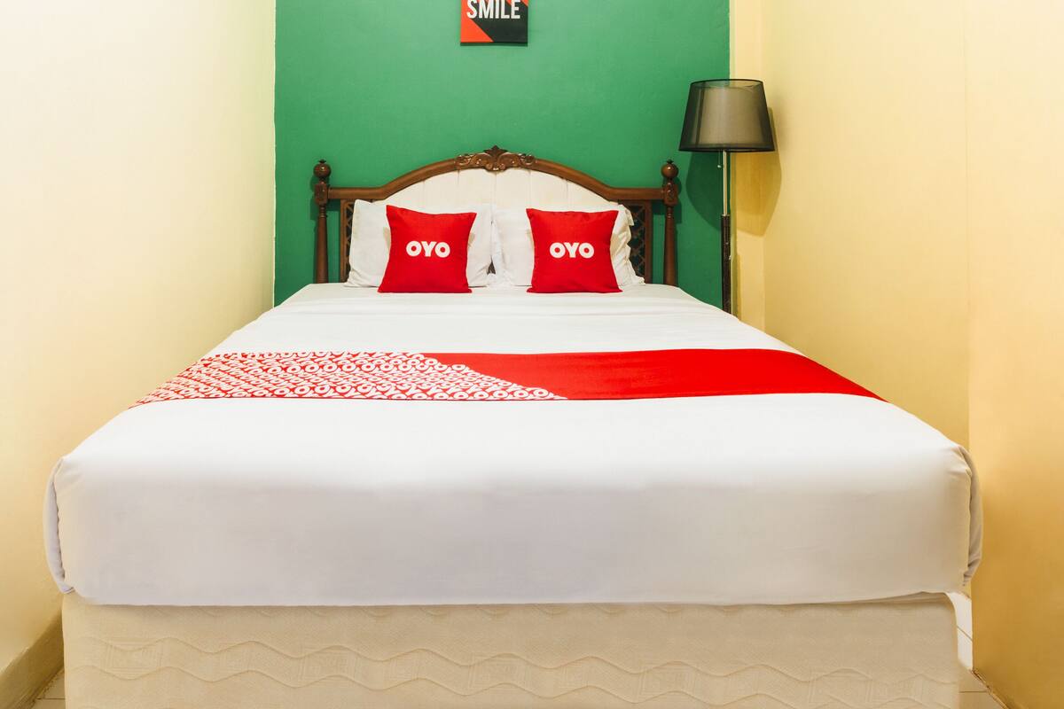 Ein Bett mit weißroter Bettdecke, zwei rote Kissen mit "OYO"-Aufschrift, ein Holzkopfstück und eine grüne Wand mit einem "SMILE"-Schild.