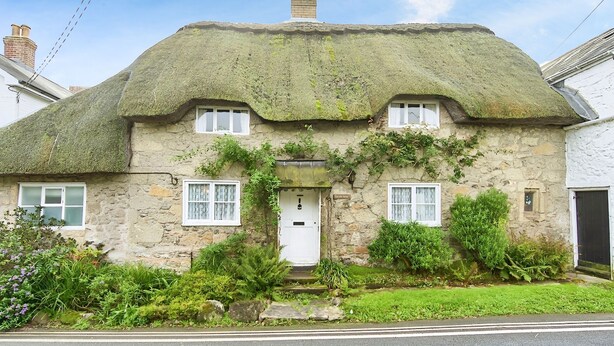 A stone house with a thatched roof, ivy-covered walls, and a white door. - Ye Olde Cottage (Ventnor)