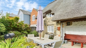 A patio with a table and chairs, a thatched-roof building, and a grill. - Ye Olde Cottage (Ventnor)