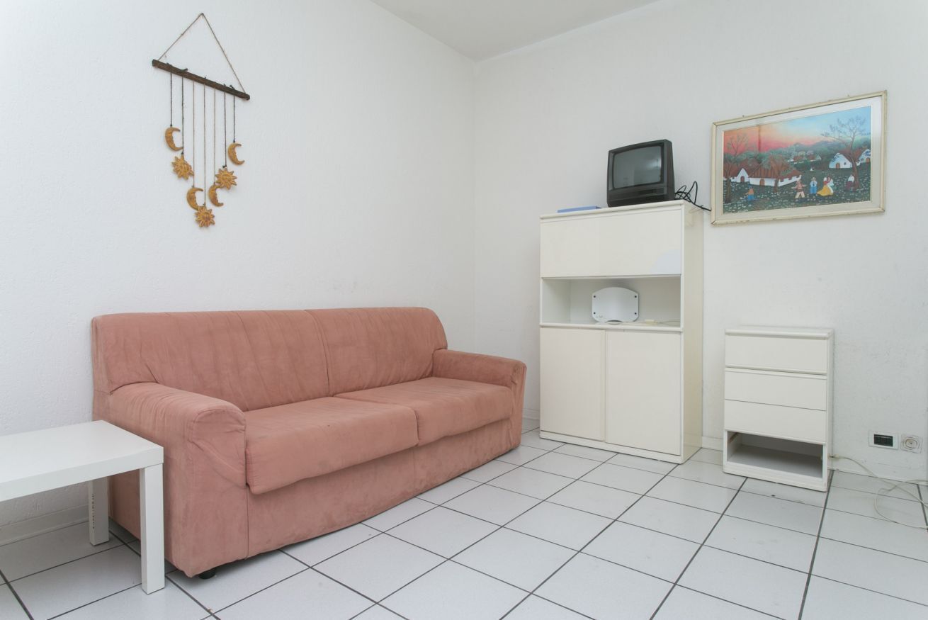 Ein Zimmer mit einem rosafarbenen Sofa, einem weißen Tisch, einem Fernseher an einer Ablage und einem Bild an der Wand.