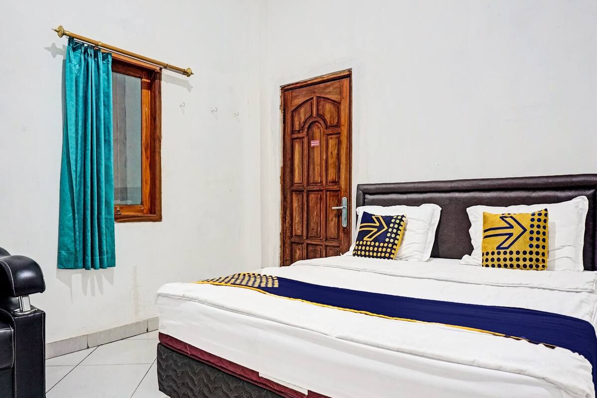 Un lit bien fait, recouvert d’une couvre-lit blanche et bleue, deux oreillers à motifs géométriques, une porte en bois et une fenêtre ornée de rideaux turquoise.