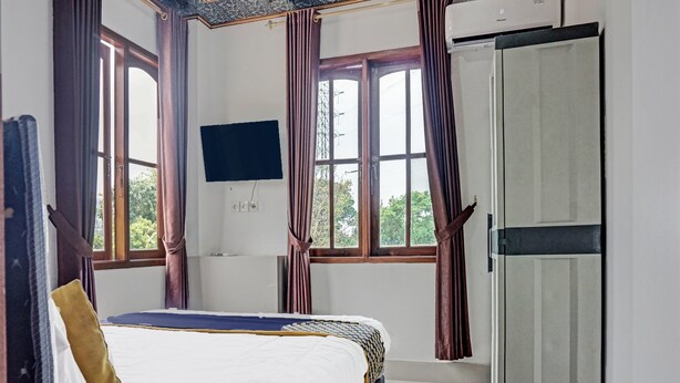 Een slaapkamer met een bed, een aan de muur bevestigde televisie, een garderobe en grote ramen met gordijnen.