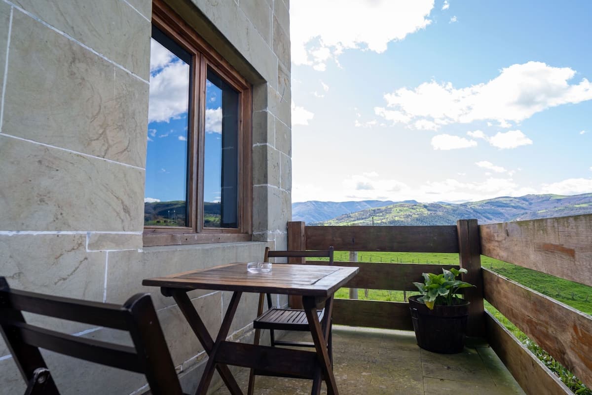 A balcony with a wooden table and chairs, a potted plant, and a view of green hills and mountains.