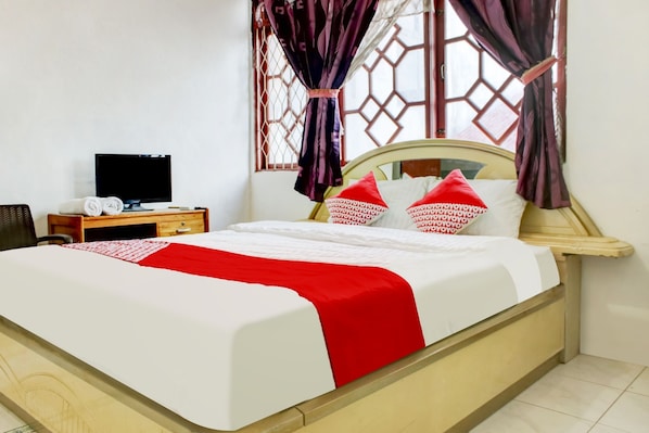 A neatly made bed with red and white bedding, a television on a wooden nightstand, and a window with decorative curtains. - OYO 2301 Hocky Guest House (Banjarmasin)