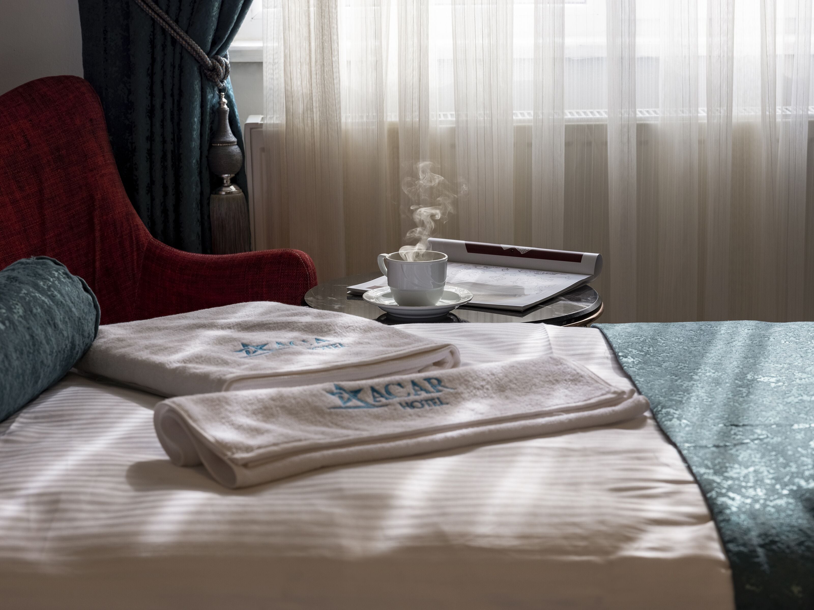 Habitación de hotel con cama, una silla roja, una mesa con una taza de café y una ventana con cortinas translúcidas.