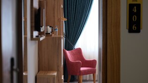 A hotel room with a wooden wardrobe, a red armchair, a small table with a kettle, and a window with curtains.