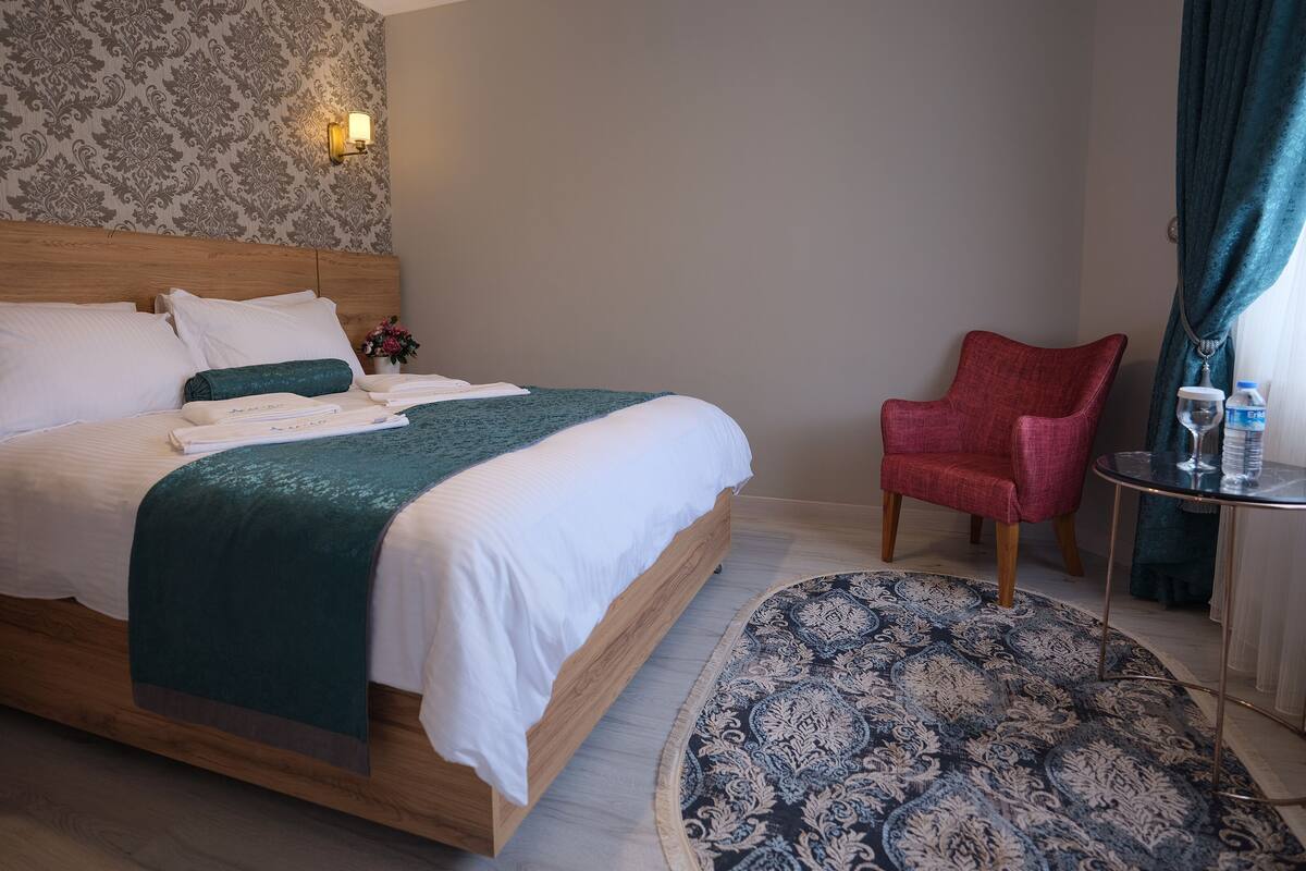 A neatly made bed with a green blanket, a red chair, a small table with a glass and water bottle, and a patterned rug.