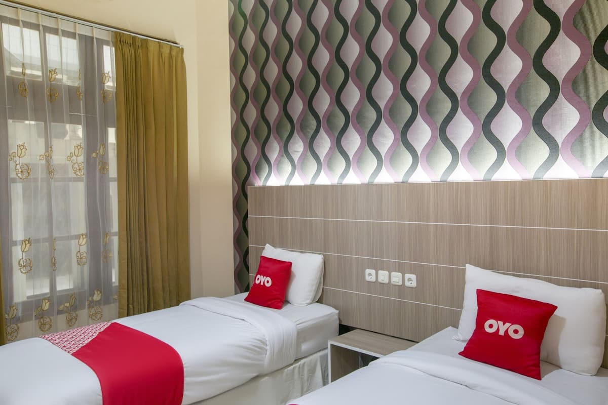 Habitación de hotel con dos camas, cada una con ropa blanca y almohadas rojas, cabeceros de madera y un papel tapiz estampado.