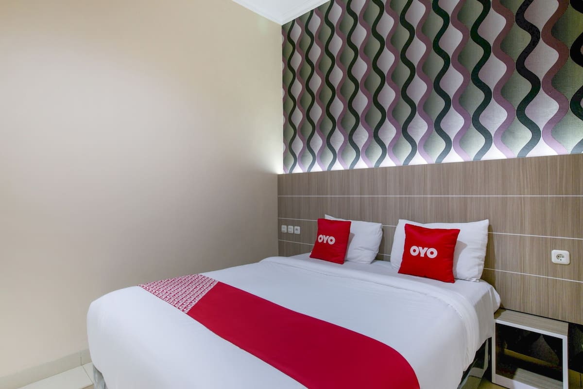 Una habitación de hotel con una cama bien hecha, dos almohadas rojas con la palabra "OYO" y una manta roja, además de un papel tapiz con diseño en la pared sobre la cama.