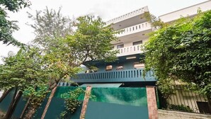 A multi-story building with a balcony, surrounded by greenery and a green fence. - Hello Stay Green (Noida)