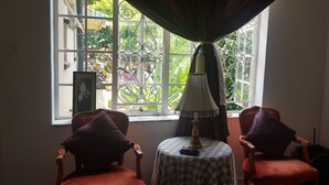 Quarto com duas cadeiras voltadas para uma janela com grade de ferro decorativa. Uma mesa com luminária e pequeno objeto decorativo fica em frente à janela.