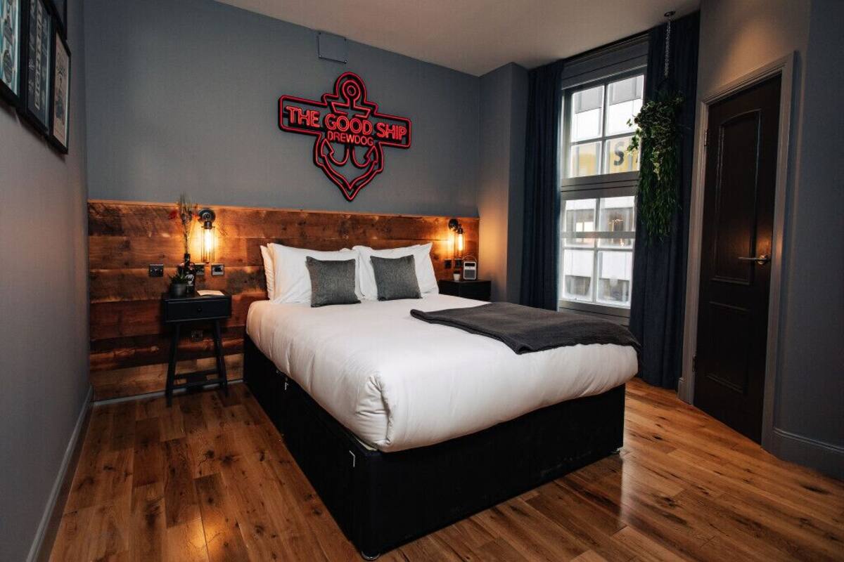 A modern hotel room with a large bed, wooden headboard, and a neon sign reading "The Good Ship Drewdog."