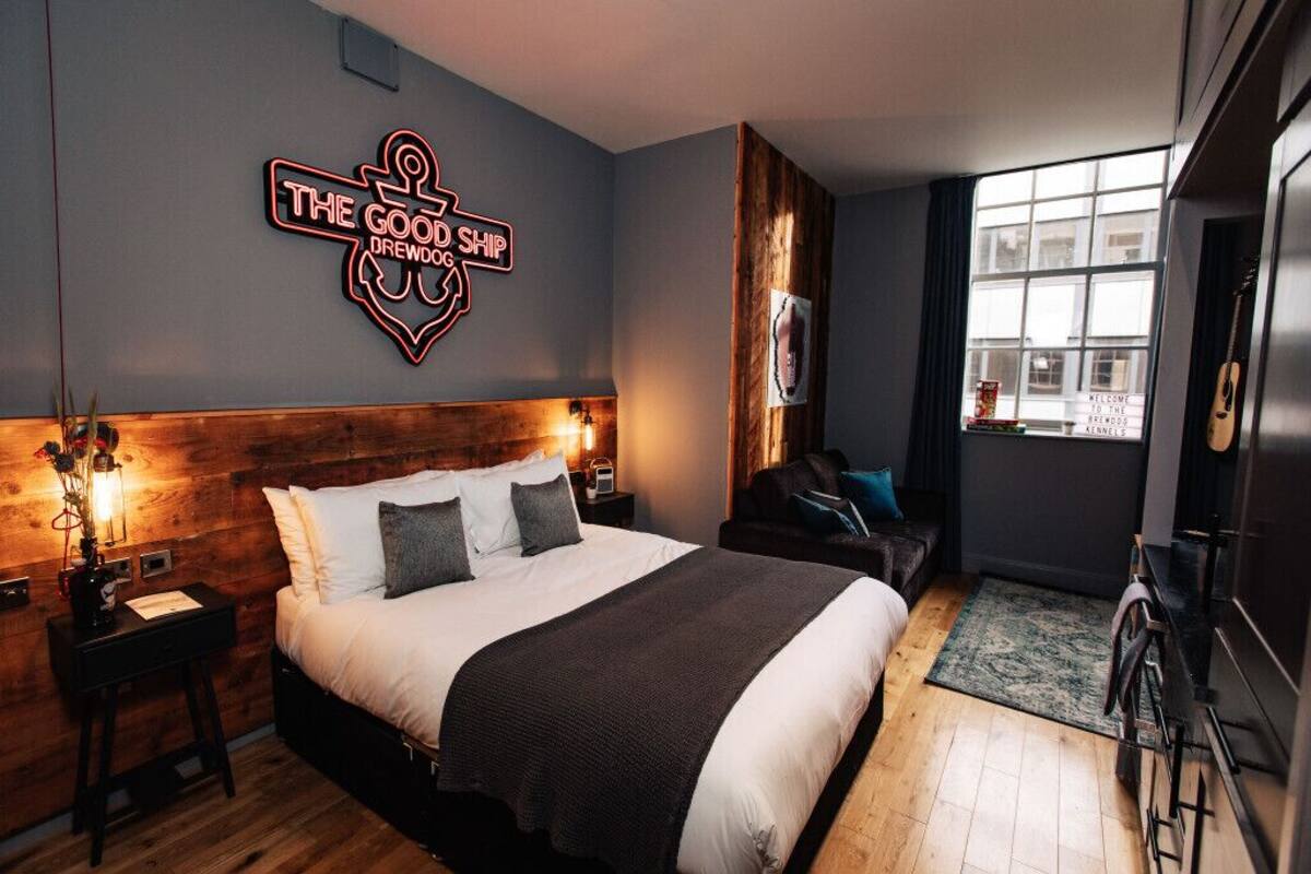 A modern hotel room with a large bed, a wooden headboard, a neon sign reading "The Good Ship Brewdog," a small seating area with a chair and a table, and a window with a view of the city.