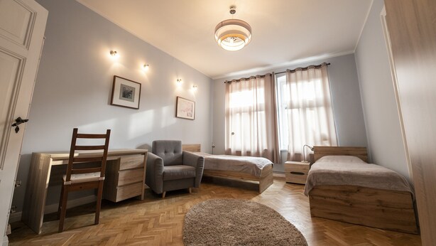 Une chambre avec un bureau en bois, deux lits simples, une chaise et une grande fenêtre avec des rideaux.