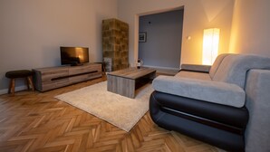 Un salon moderne avec un parquet en bois disposé en chevrons, un canapé gris, une table basse en bois, un téléviseur posé sur un meuble en bois et une cheminée en pierre.