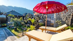 A serene outdoor area with lounge chairs and a red umbrella, surrounded by a natural rock garden and a traditional building. - Latengaya Mount Batur Bungalow (Kintamani)