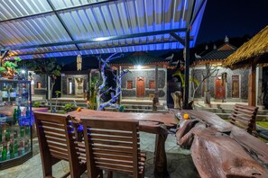 Un área de estar al aire libre con mesas y sillas de madera, una vitrina de vidrio con botellas y un muro de piedra con puertas de madera.