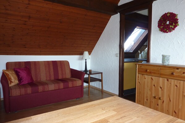A cozy room with a wooden ceiling, a red sofa with patterned cushions, a small table with a lamp, and a wooden cabinet.