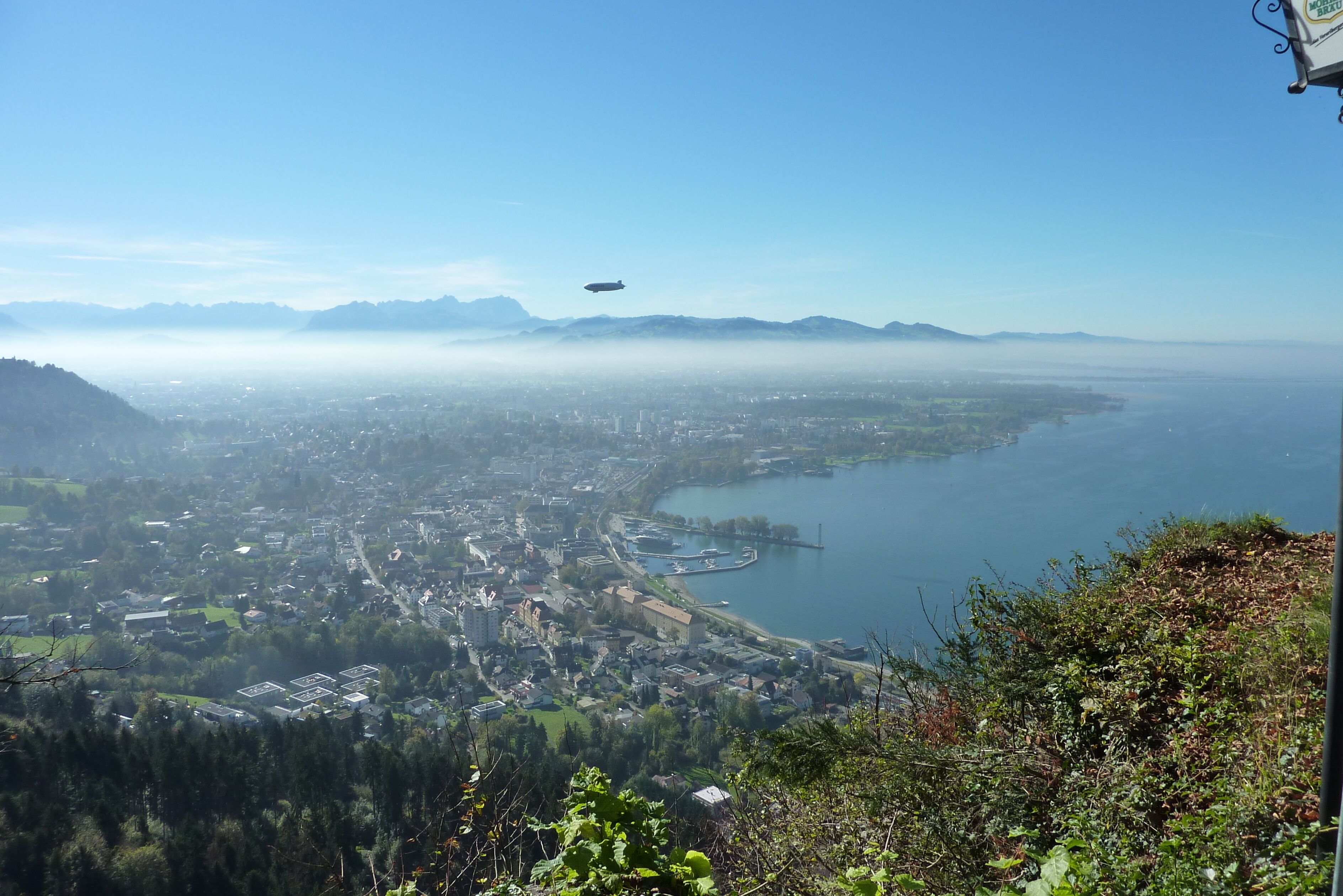 Vista panorâmica de uma cidade costeira, com um helicóptero sobrevoando, cercada por montanhas e um corpo d'água.