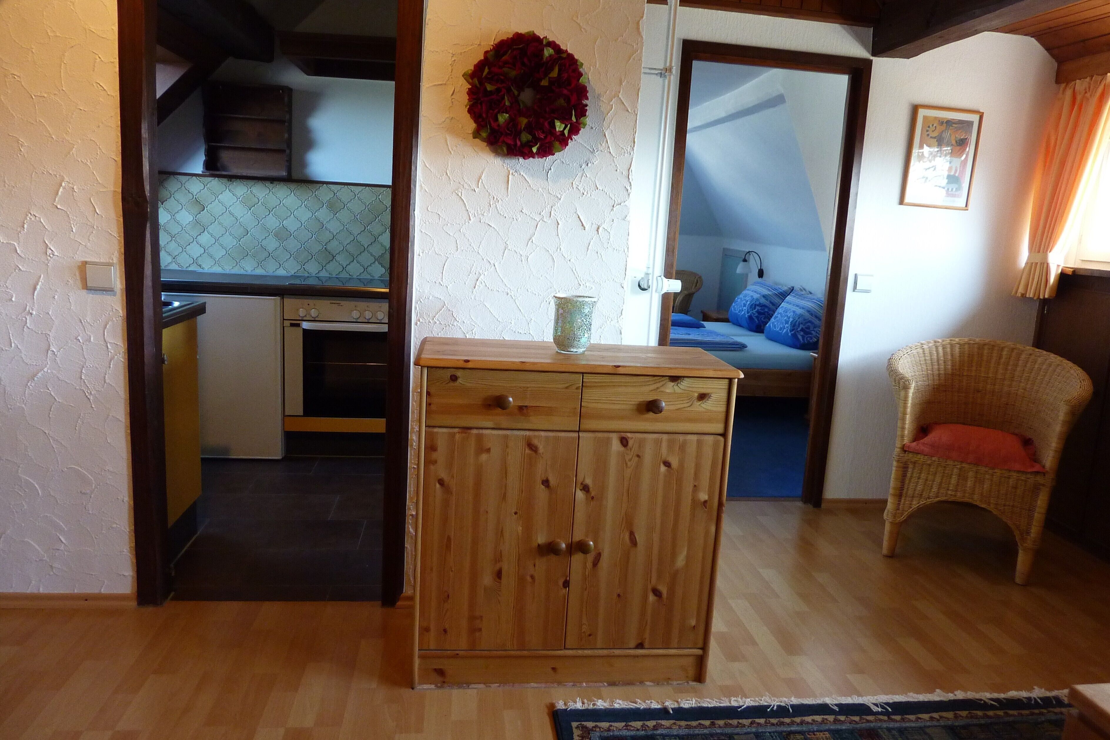 A room with a wooden cabinet, a wicker chair, and a bed with blue pillows.
