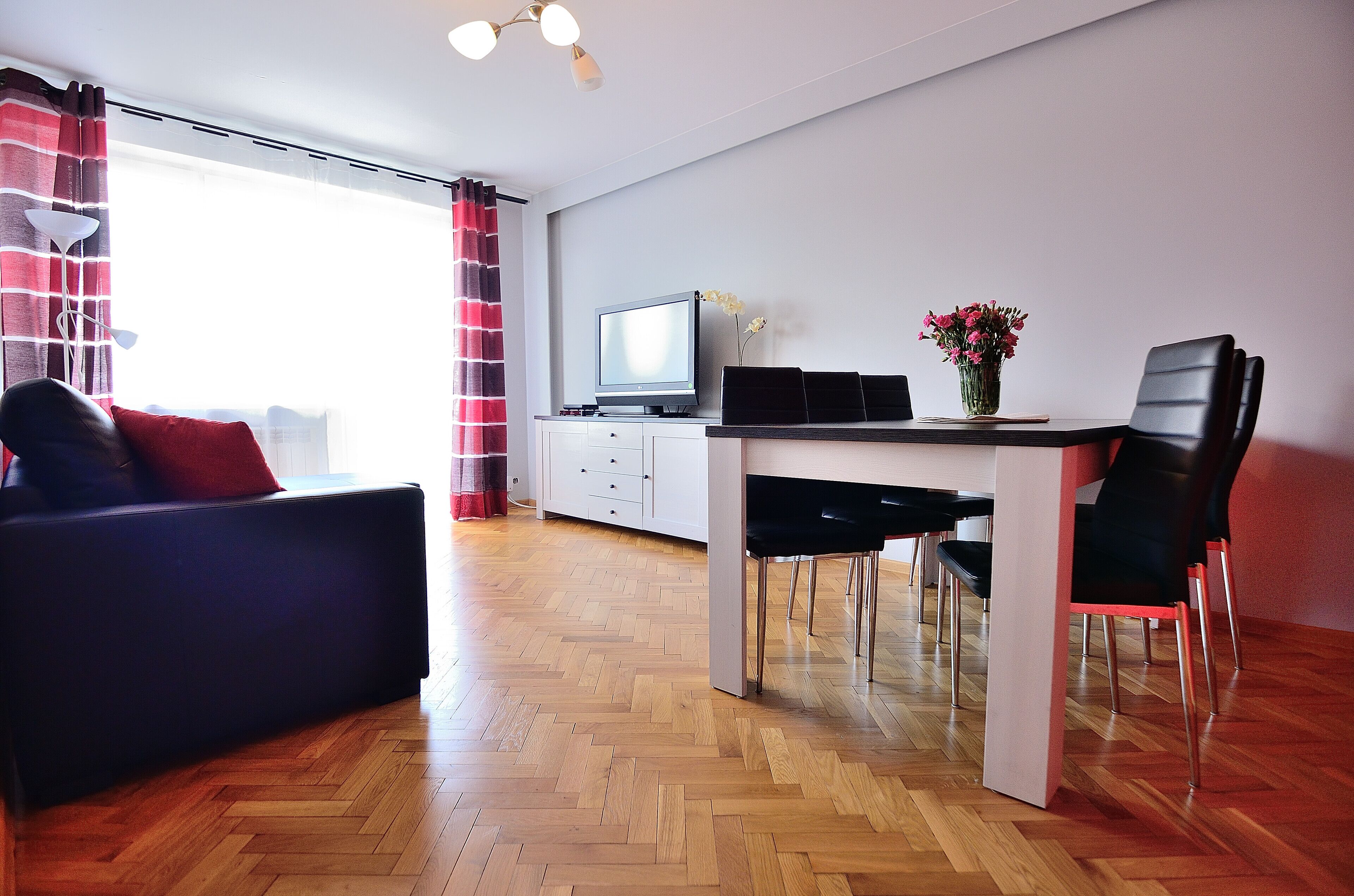 A modern living room with a sofa, a dining table with chairs, a TV, and a vase with flowers.