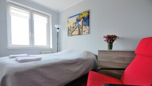 A bedroom with a bed, a red chair, a dresser, a painting, and a vase with flowers. - Victus Apartmenty - Jantar (Sopot)