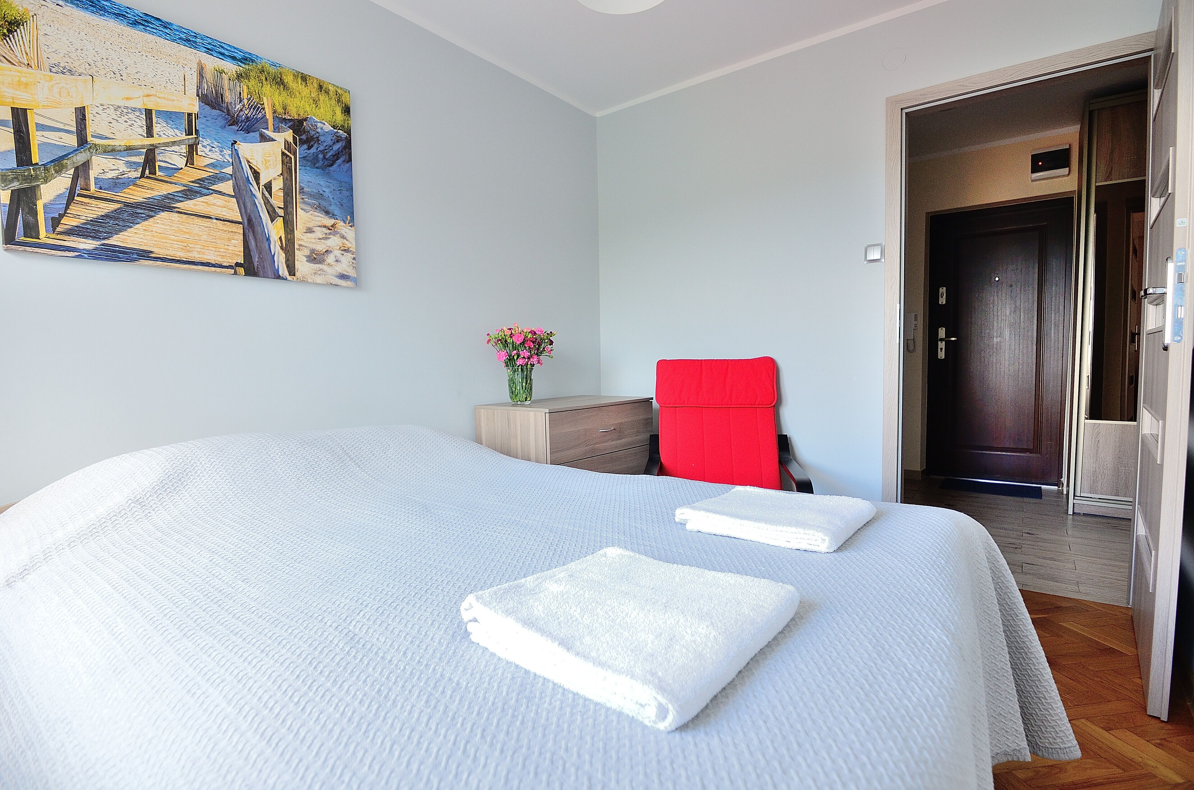 A hotel room with a large bed, a painting of a beach scene, a wooden nightstand, a red chair, and a door leading to another room.