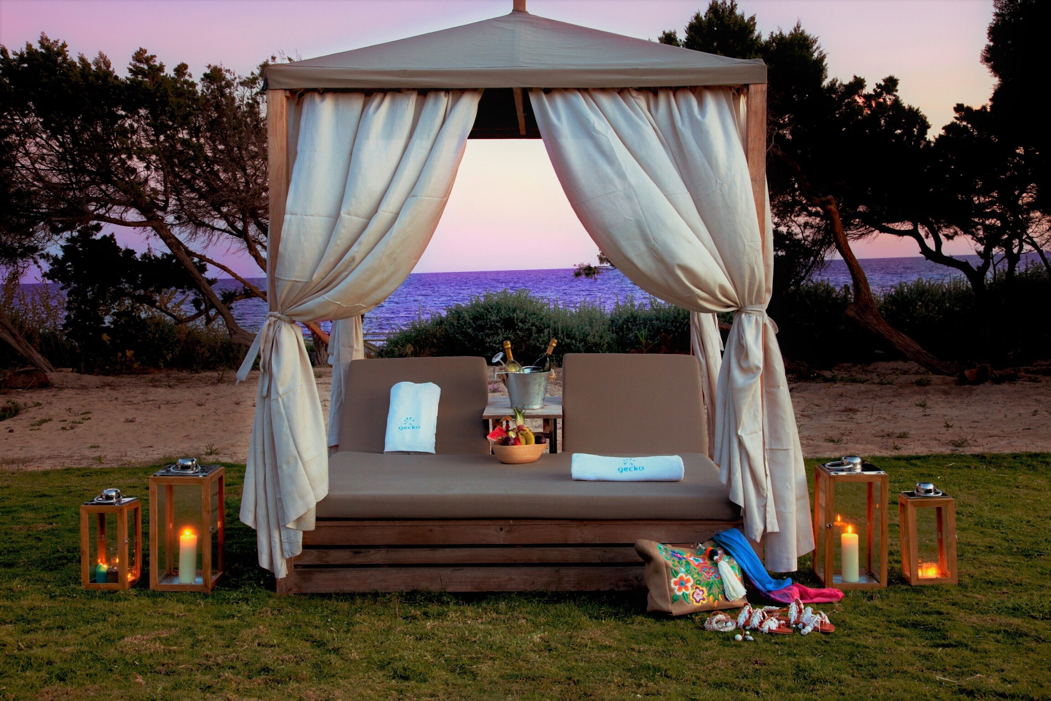 A beachside gazebo with a cushioned seating area, a bowl of fruit, and candles.